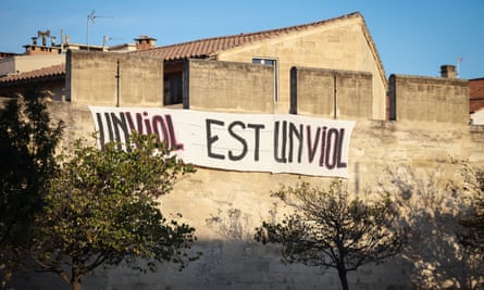 A banner reads ‘rape is rape’; it is tied high on a stone wall above trees with rooftops in the background and a clear blue sky