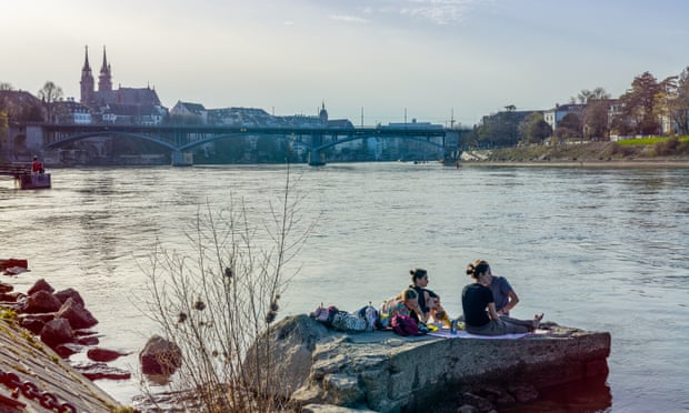 People sit on a concrete slab at the edge of the River Rhine in Basel with a view of the city skyline. Switzerland.