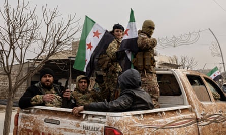 Five fighters – two standing and holding opposition flags, three seated – ride in the back of a Toyota pickup truck. They wear army fatigues and several have rifles; one wears a balaclava