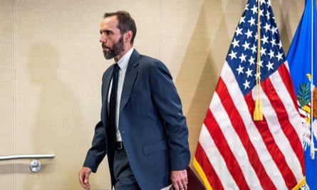 A white, thin man with dark hair a full, scruffy dark beard in a blue suit and tie, walks past an American flag holding something like a file folder.