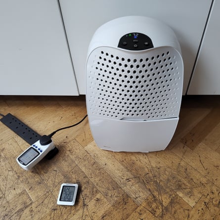 Ebac dehumidifier on a wooden floor. Next to it is a power meter plugged into a black multi-way plug, and a humidity testing device