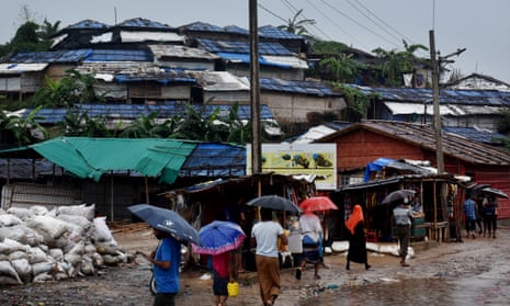 People holding umbrellas walk along a road next to piled-up sandbags and makeshift buildings