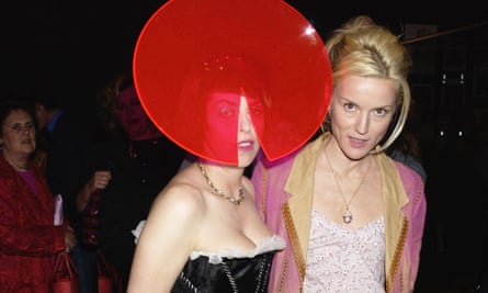 Daphne Guiness stands next to Isabella Blow who is wearing a hat with a large red circle slanted over her face, and a segment cut out over her mouth and nose