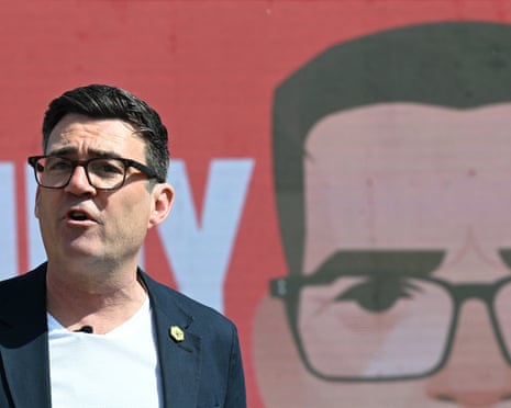 Andy Burnham, in a T-shirt and blazer, speaks in front of a billboard showing a close-up cartoon image of his face and oblong-framed glasses