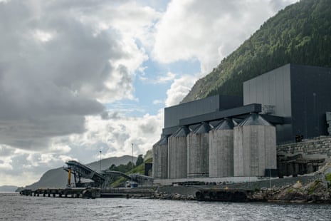 A mining plant beside a body of water against a steep tree-covered mountainside