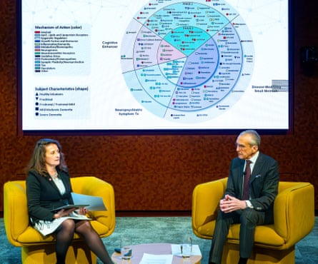 A middle-aged white woman and an older man sitting on stage in armchairs with a big display behind them.