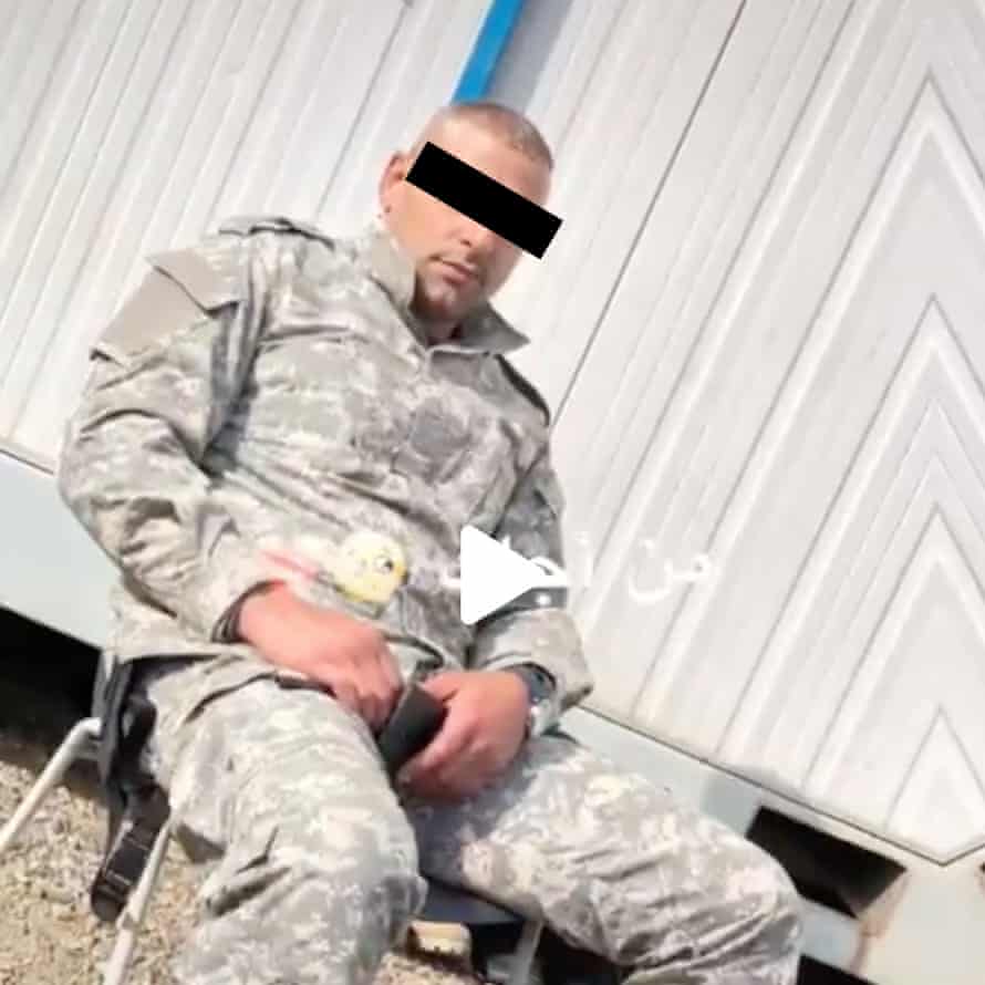 A seated man in faded military fatigues with what looks like a handgun by his side and his eyes covered to protect his identity  