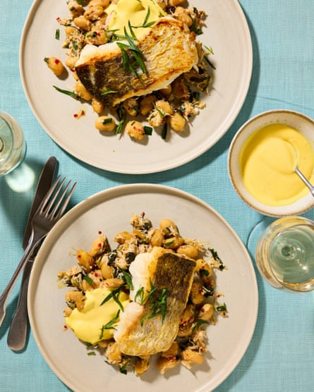 Two plates of the roasted fish alongside a bowl of the aioli and cutlery and drinks, served on a blue tablecloth. Andy Beynon’s roast hake with saffron aioli and crabby anchovy roasted garlic butter beans with tarragon.
