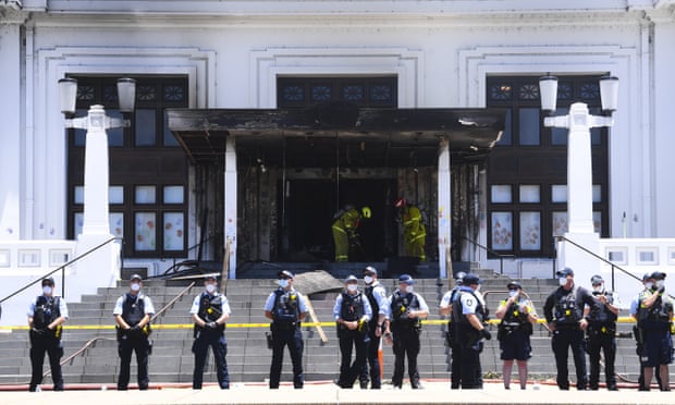 A line of police officers stands in front of the steps to the doors of old Parliament House in Canberra. Two firefighters are standing in front of the doors, which are badly damaged and charred by fire.