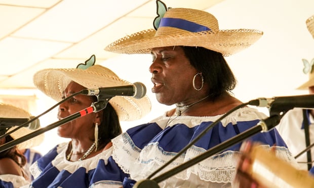 Woman wearing a white and blue ruffled dress and straw hat sings into a mike.