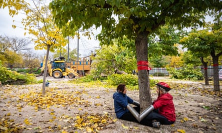 Two women, one in a bike helmet, sit on the ground on either side of a tree, their arms joined together while inserted into V-shaped metal pipes