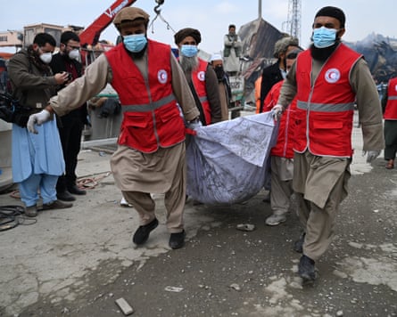 Four Afghan Red Crescent Society volunteers wearing red tabards and facemasks carry victims’ bodies in a sheet.