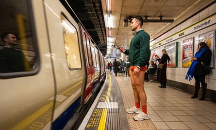 A man stands on a London Underground platform in a green jersey, orange pants and socks and white trainers