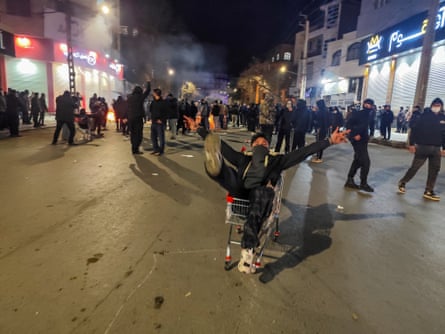 A man sits in a shopping trolley in the middle of a road. Around him, people in dark clothes, many covering their faces with masks, walk around the street