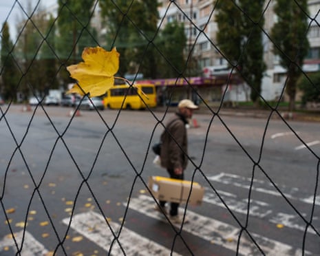 Seen through an anti-drone net with a yellow leaf caught in it, a man carrying a box crosses an Oleshky street
