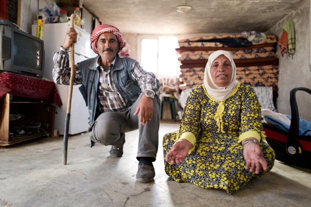 An older couple look at the camera in their home – the woman sitting and the man squatting while holding on to a stick