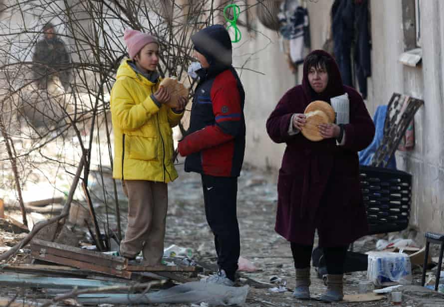 Female resident holding loaves of bread outside a damaged building in Mariupol, 28 March. On left of picture, a child eats from a loaf of bread.