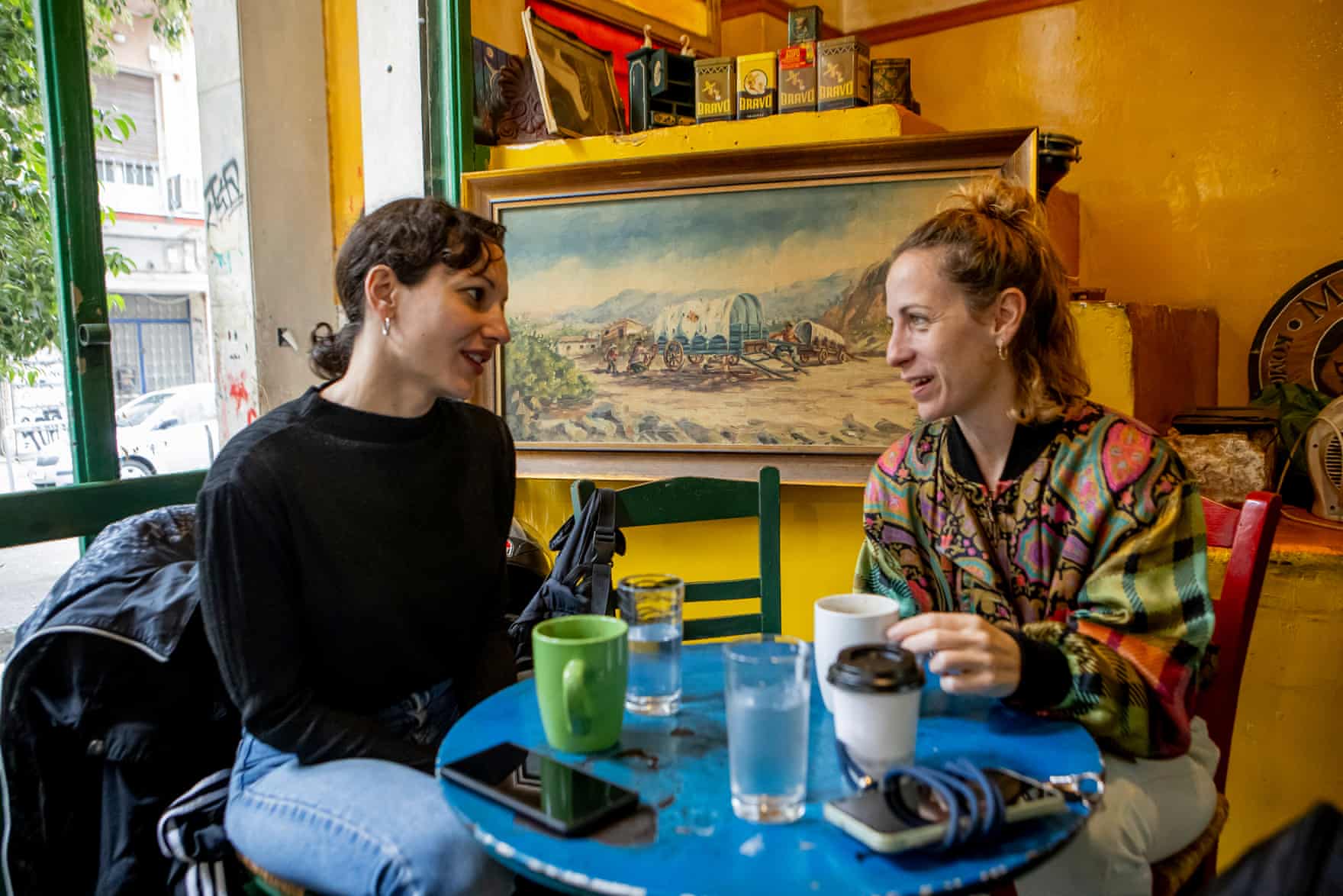 Two women talk over mugs of coffee at a blue circular table with a painting behind them. They are quite young, one dark-haired and the other blond. The blond woman wears a brightly patterned zip-up jacket.
