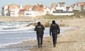 Two police officers walk along the water line of a stony beach with houses in the background