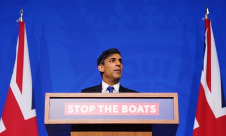 Rishi Sunak, in a shirt, tie and suit jacket, looks right as he speaks at a podium with a “Stop the boats” sign and a Union Jack flag on either side of him