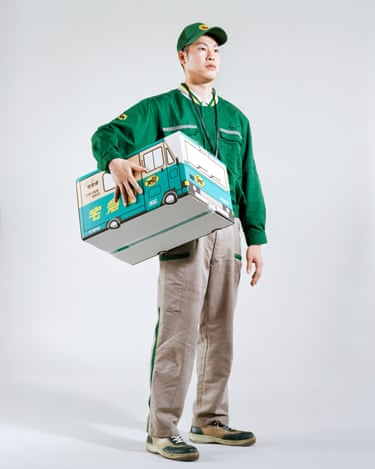 A delivery man in green uniform carries a cardboard box shaped like a delivery truck