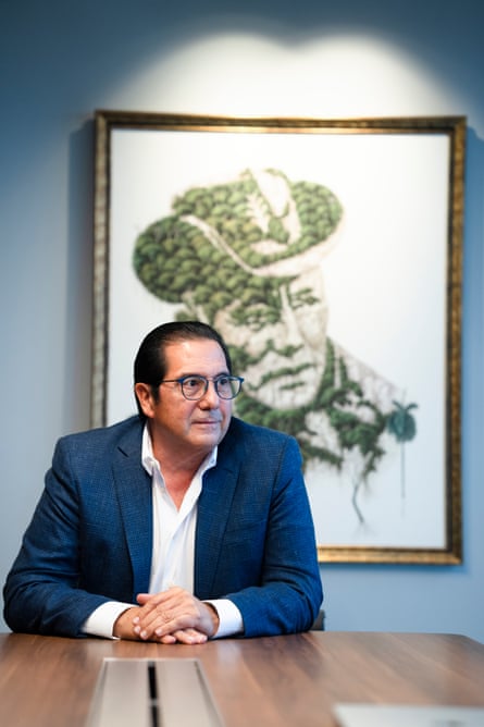 Martin Torrijos, in a shirt, jacket and glasses, seated at a wooden table with his hands folded in front of a portait of his father wearing a folded-brim hat