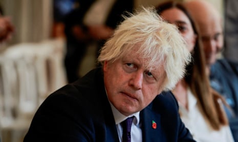 Boris Johnson sitting and looking up an someone who is speaking wearing a dark jacket and a poppy badge.