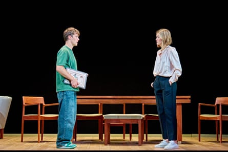 A young man (actor Jasper Talbot) and middle-aged woman (Rosamund Pike) stand on stage facing each other, as if talking, looking serious, with a mid-century style dining table and chairs behind them