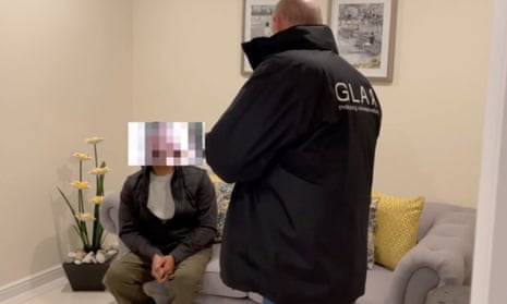 A Gangmasters and Labour Abuse Authority agent standing above a suspect, who is sitting on a sofa and has their face pixellated