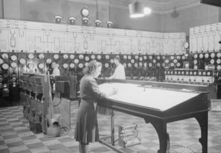 A general view of the control room at a British power station, 1945. A woman stands at a tall, illuminated desk in a room surrounded by control panels and circuitry.