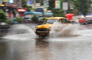 A yellow 1950s taxi sends up a cloud of spray as it drives along a flooded street
