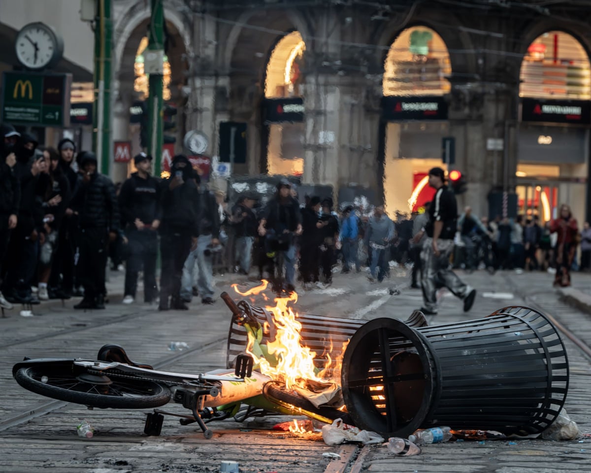A burning bicycle and bin sit in a Milan street during a demonstration, crowds gathered nearby