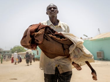 A man carries his malnourished daughter in blankets across a dusty yard