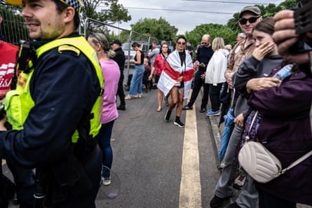 Protesters outside the Bell hotel; a police officer stands in front of them and they are all behind metal fencing. One woman has an England flag over her shoulders.