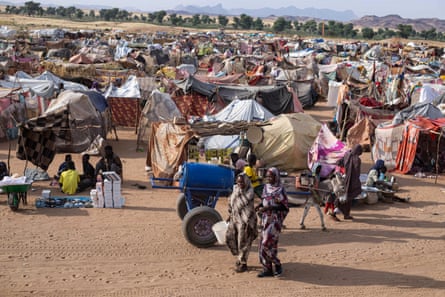View across camp with two women walking in foreground, one carrying a plastic bucket; behind them is a donkey and cart, and a stall with goods including sandals laid out; lines of makeshift shelters made from sticks and coloured blankets and sheets stretch across the barren, sandy landscape