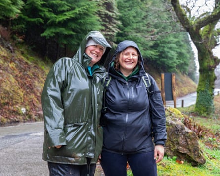 Two women wearing raincoats with their hoods up standing side by side on a country path and smiling.