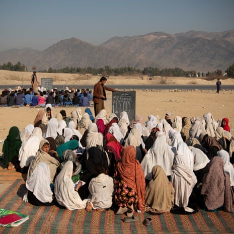 A crowd of girls huddle on the ground in front of a man with a blackboard; a similar huddle of boys can be seen in the distance. The terrain is dusty, mountains can be seen in the background