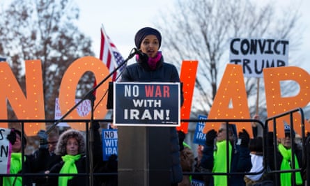 Ilhan Omar on a podium, addressing a rally, with a sign in front of her saying 'No war with Iran!' and big orange letters behind her spelling out 'No war'