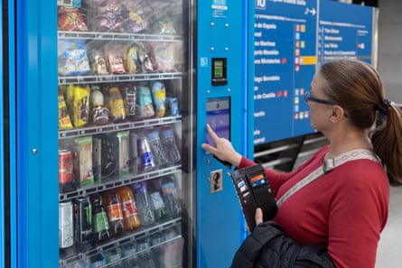 Mature woman using a blue touchscreen vending machine at a subway station, selecting snacks and drinks and getting ready to pay with a card.