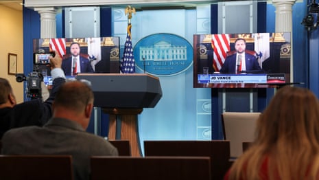man in suit speaks at lectern with finger pointed, an american flag behind him