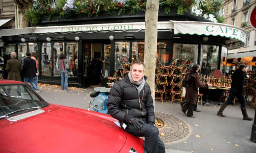 Vallee in a coat sitting on the bonnet of a red car with the picturesque Cafe de Flore itself behind him, with all its outside chairs in stacks