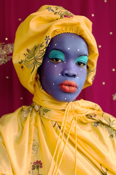 A woman with blue-painted face and teal eyeshadow wears an ornate yellow headwrap against a magenta backdrop