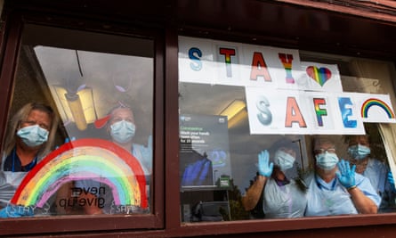 Staff in masks and gloves looking out of a window at the photographer. A painted rainbow and ‘Stay safe” posters are stuck to the window, as well as a government poster about Covid.