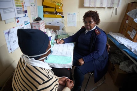 A woman in a medical uniform sits facing a patient wearing a face mask in a small clinic room.