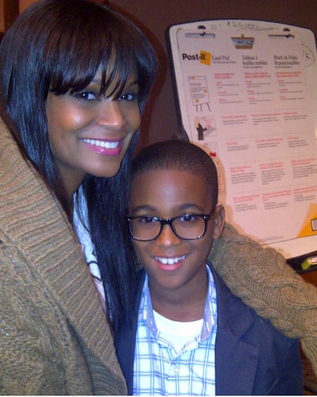 A young boy wearing glasses smiles next to his mother