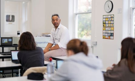 Steve Green takes a GCSE biology class – he is seated in a classroom facing three girls whom we see from behind; he wears an open white shirt and pale jeans, one of the girls wears a hoodie, and a relaxed atmosphere is suggested