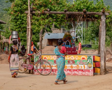 Two women, one carrying a metal container on her head, walk past a colourfully painted roadside structure