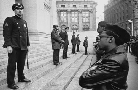 A line of Black Panthers stand with arms crossed facing a line of New York City police officers standing on a few steps above them.