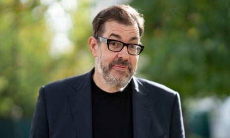 Richard Osman – he is standing outdoors with light shining through blurred trees in background, and is wearing a dark jacket, black jumper and black-rimmed glasses; he has a short greying beard and swept-back light brown hair