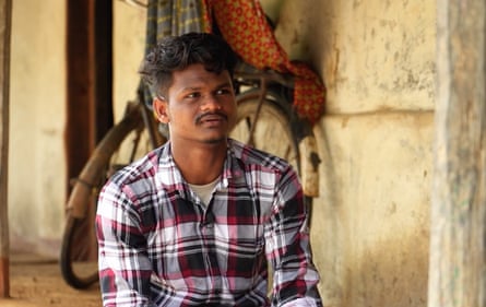 A young man in a plaid shirt sits in front of the wall of a house.